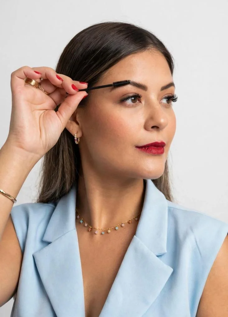 Amanda Farias, de blazer azul-claro, penteando a sobrancelha com uma escovinha.